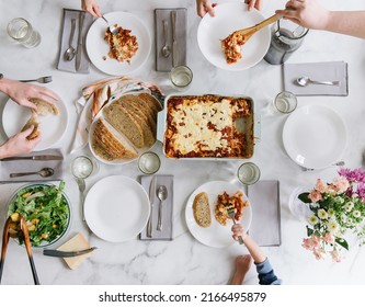 A Family Of 6 Is Sitting At The Table Serving Lasagna, Homemade Bread, And Salad. There Are Also Some Flowers To The Side Of The Table. Dinner Is Served. 