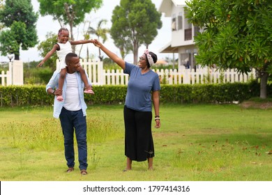 Family Of African American People With Young Little Daughter Walking On Green Grass Field While Enjoying Summer Garden Outside The House In The Neighborhood With Copy Space