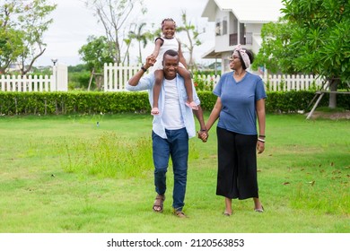 Family Of African American People With Young Little Daughter Walking On Green Grass Field While Enjoying Summer Garden Outside The House In The Neighborhood