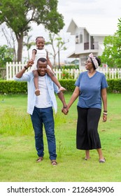 Family Of African American People With Young Little Daughter Walking On Green Grass Field While Enjoying Summer Garden Outside The House In The Neighborhood