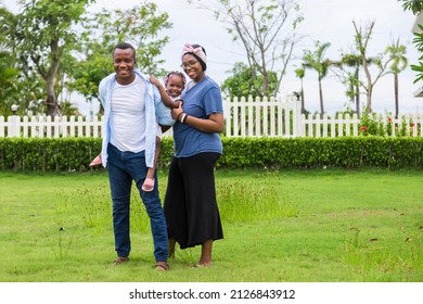 Family Of African American People With Young Little Daughter Walking On Green Grass Field While Enjoying Summer Garden Outside The House In The Neighborhood