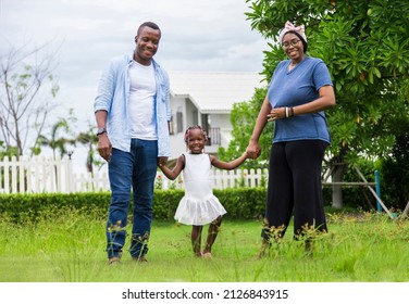 Family Of African American People With Young Little Daughter Walking On Green Grass Field While Enjoying Summer Garden Outside The House In The Neighborhood