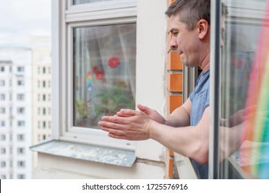 Family Applauding Medical Staff From Their Balcony. People Clapping On Balconies And Windows In Support Of Health Workers During The Coronavirus Pandemic. A Rainbow Is Drawn On The Window.