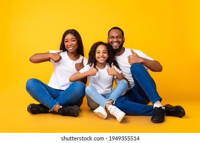 Family Approval. Portrait Of Smiling Black Family Showing Thumbs Up Sign Gesture With Both Hands, Recommending Something Cool. Mom, Dad And Daughter Sitting On The Floor At Yellow Studio