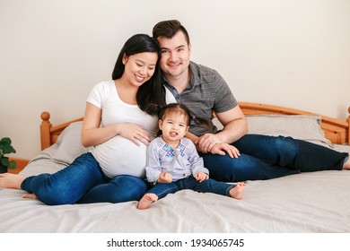 Family Asian Chinese Pregnant Woman And Caucasian Man With Mixed Race Toddler Girl Sitting On Bed At Home. Mother, Father, Baby Daughter Expecting A New Family Member. Ethnic Diversity. 