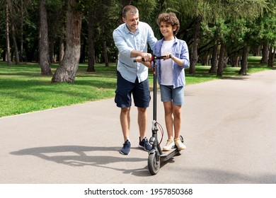 Family Assistance. Full Body Length Portrait Of Smiling Adult Father Teaching His Son Cycling At Public Park, Pushing E-scooter, Helping Him Driving And Holding Handlebar, Happy Boy Learning To Drive