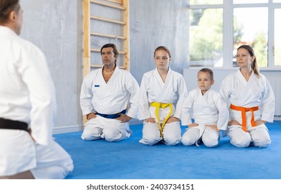 Family of athletes sit in row in front of man coach and listen attentively. Sports mentor communicates with group of Aikido students before training.