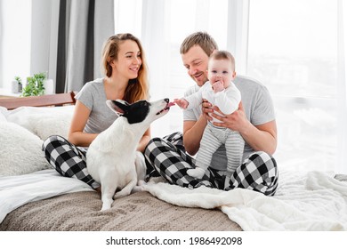 Family With Baby Boy Sitting On The Bed With Cute Dog. Mother And Father With Their Son And Doggy Together Smiling. Beautiful Parenthood Time. Pet With Owners