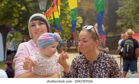 Family With Baby. Happy Couple With A Child Walk Next To Rainbow Flags, Hugs, Smiling. To Change Wider Society Attitude Towards Respect, Full Acceptance, And Inclusion Of LGBT People In Society.