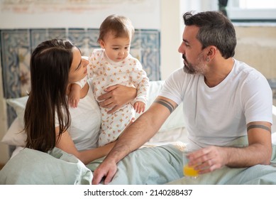 Family With Baby Sitting Together In Bed In Morning. Mid Adult Woman Holding Little Son And Man Drinking Juice. Family At Home Concept