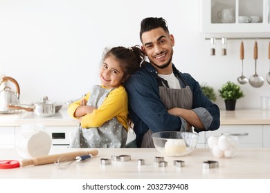 Family Baking. Portrait Of Happy Arab Dad And Little Daughter In Aprons Posing In Kitchen, Middle Eastern Father And His Cute Female Child Standing Back To Back, Ready For Cooking Pastry Together