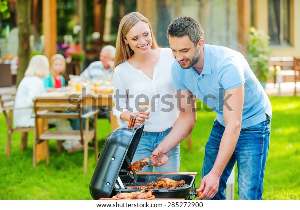 Family barbeque. Happy young couple\
barbecuing meat on the grill while other members of family sitting\
at the dining table in the background\

