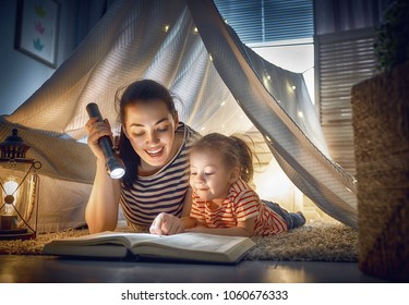Family bedtime. Mom and child daughter are reading a book in tent. Pretty young mother and lovely girl having fun in children room.