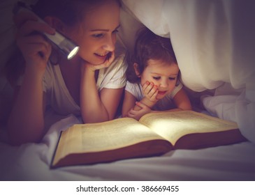 Family Bedtime. Mom And Child Daughter Reading A Book With A Flashlight Under The Blanket In Bed