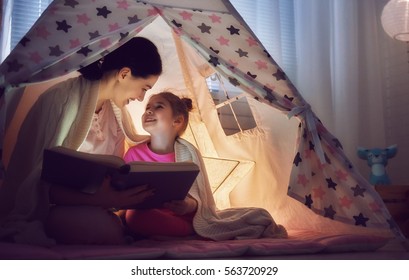 Family Bedtime. Mom And Child Daughter Are Reading A Book With Flashlights In Tent. Pretty Young Mother And Lovely Girl Having Fun In Children Room.