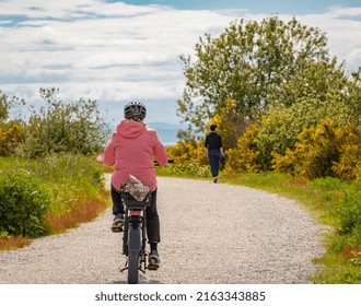 Family Bike Ride In Sunny Spring Day Along Fields And Forest. An Elderly Woman Riding On A Park Road. People Cycling On Trail Between The High Grass On An Embankment-May 29,2022-Delta BC Canada