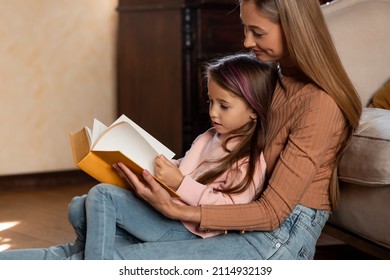 Family Bonding Concept. Caring Young Woman Reading Book Aloud To Her Cute Little Daughter, Happy Mommy And Kid Enjoying Spending Time Together At Home, Sitting On Floor Leaning On Sofa In Bedroom