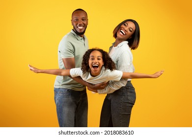 Family Bonding Concept. Joyful Black Man And Woman Holding Daughter On Hands, Having Fun Over Yellow Studio Wall. Smiling Girl Pretending She Is Flying, Spreading Hands, Imitating Plane