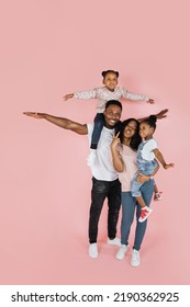 Family Bonding Concept. Joyful Black Man And Woman Holding Daughters On Hands, Having Fun Over Pink Studio Wall. Smiling Girls Pretending They Are Flying, Spreading Hands, Imitating Plane