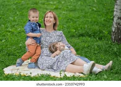 Family bonding outdoors: A mother and her two children share a tender embrace while seated on a picnic blanket in a serene park setting. - Shutterstock ID 2310742993