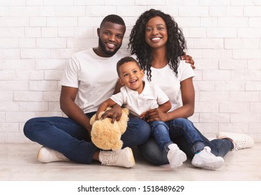 Family Bonds. Young African American Parents And Son Sitting On Floor Together, Looking At Camera Over White Brick Wall Background, Copy Space