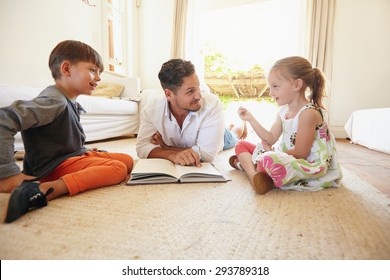 Family With Book Sitting On Floor With Little Girl Talking To His Father And Brother. Father With His Little Boy And Girl At Home.