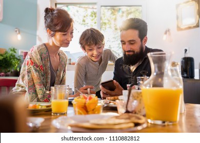 Family Breakfast, Dad Shares A Video On A Phone To His Son. Mom Looks Over Her Son's Shoulder As The Morning Sun Enters Through The Window