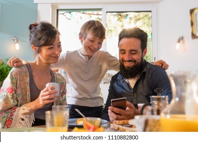 Family Breakfast, The Father Shows Pictures On His Phone To His Wife And Son. Everyone's Having Fun While The Morning Sun Comes In Through The Window