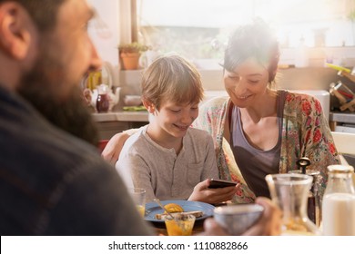 Family Breakfast In The Kitchen, A Boy Shows Pictures On His Phone To His Mother. Everyone's Having Fun While The Morning Sun Comes In Through The Window