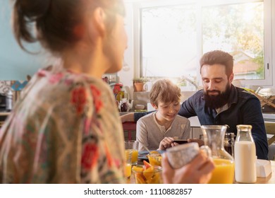 Family Breakfast In The Kitchen, The Father Shows Pictures On His Phone To His Son. Everyone's Having Fun While The Morning Sun Comes In Through The Window