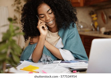 Family Budget And Finances. Cute African Woman With Afro Haircut And Braces Having Phone Conversation And Smiling While Doing Paperwork, Calculating Domestic Expenses, Paying Bills Online On Laptop