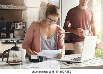 Family budget and finances. Young woman doing accounts together with her husband at home, planning new purchase. Serious female in glasses holding piece of paper and making necessary calculations