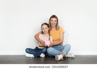 Family Budget. Smiling Beautiful Mother And Little Daughter Holding Piggy Bank While Relaxing On Floor Near White Wall, Happy Mom And Female Kid Enjoying Economy And Money Savings, Copy Space - Shutterstock ID 2311020637