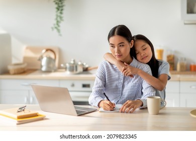 Family Business. Young Asian Woman Working On Laptop And Taking Notes, Loving Cute Daughter Embracing Mother, Spending Time Together At Home, Sitting In Kitchen Interior