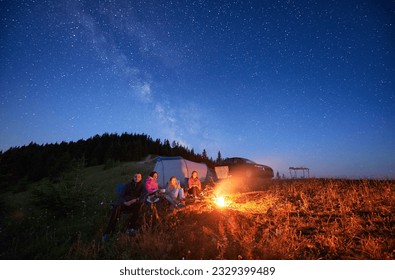 Family camp in mountains in the evening. Group of tourists having a rest near campfire on a top of hill, near tourist tent and off-road vehicle. Starry night in Carpathian mountains. - Powered by Shutterstock