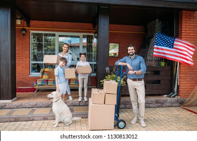 Family With Cardboard Boxes And Labrador Dog Standing On Country House Porch, Moving Home Concept