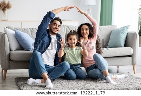 Family care. Arabic parents making symbolic roof of hands above cute little daughter while sitting together on floor in living room, middle eastern mom and dad having fun with their child at home