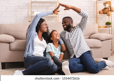 Family Care And Protection Concept. Black Parents Making Symbolic Roof Of Hands Above Their Little Daughter, Sitting On Floor At Home