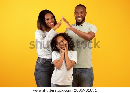 Family care, protection and insurance concept. Young black parents making symbolic roof of hands above little daughter while standing over yellow background, studio shot