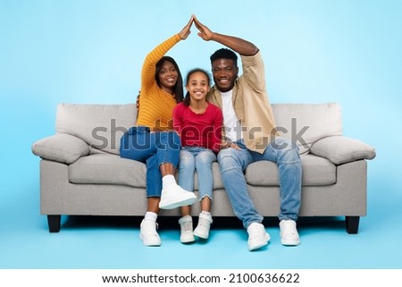 Family Care, Protection And Insurance Concept. Portrait of smiling young African American parents making symbolic roof of hands above their happy daughter sitting on sofa couch at blue studio wall