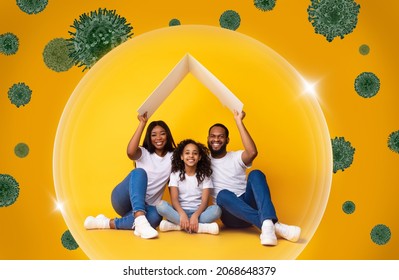 Family Care And Protection While Coronavirus Pandemic Concept. Smiling Black Parents Sitting Under Symbolic Cardboard Roof Above Them And Happy Daughter, Three People Isolated From COVID-19 Bacteria