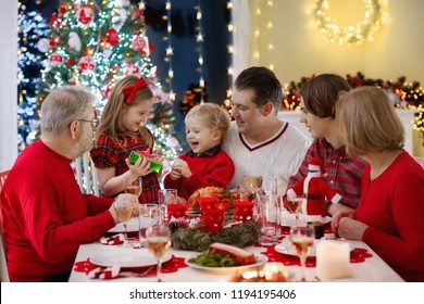 Family with children eating Christmas dinner at fireplace and decorated Xmas tree. Parents, grandparents and kids at festive meal. Winter holidays celebration and food. Kids open presents and gifts.