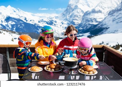 Family With Children Enjoying Apres Ski Lunch With Traditional Swiss Raclette And Cheese Fondue In Restaurant On Top Of Snow Covered Mountain On Winter Or Christmas Vacation. Parents And Kids Skiing.