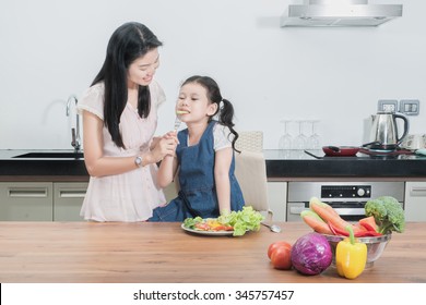 Family, Children And Happy People Concept - Asian Mother Feeding Kid Daughter Vegetables In Kitchen.