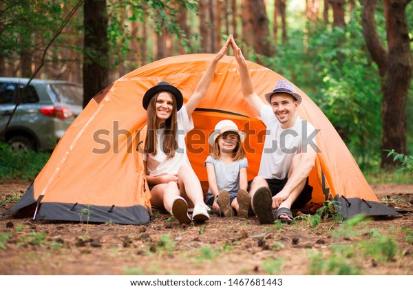 Family with children making arms roof promise\
protection forever. New building house purchase concept. Stylish\
full family with kid sitting near tent in forest camp, mom and dad\
making roof figure