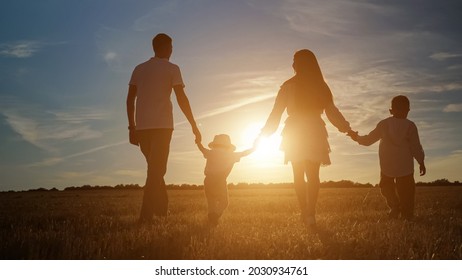 Family with children sons silhouettes walks joining hands along shadowed field at back setting sun in summer under blue sky backside view, sunlight