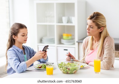 Family, Communication And People Concept - Sad Mother Looking At Her Daughter With Smartphone Having Dinner At Home