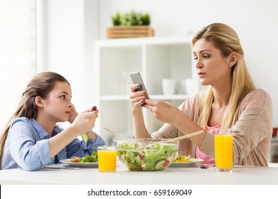 Family, Communication And People Concept - Sad Girl Looking At Her Mother With Smartphone Having Dinner At Home