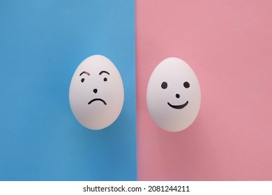 
Family Concept, No People. Eggs With Painted Emotions On A Blue And Pink Paper Background. One With A Displeased Expression, The Other With A Smiling Face. Copy Space. 