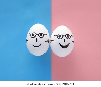 Family Concept, No People. Two Eggs With Painted Emotions On A Blue And Pink Paper Background. They Have Poor Eyesight, So They Are Both Wearing Glasses, Smiling And Holding Hands. Copy Space. 